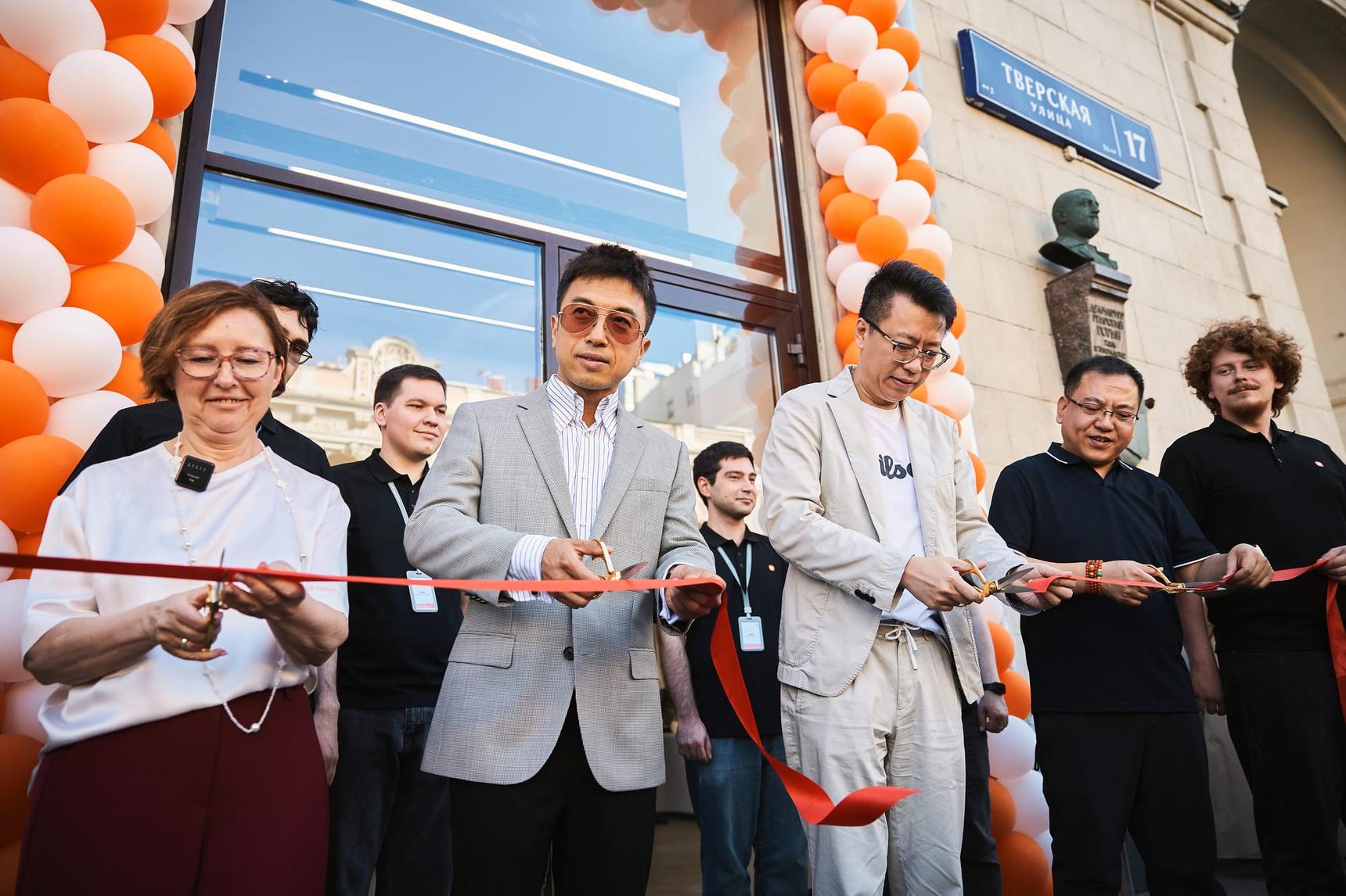 Открытие магазина Xiaomi: группа людей режет красную ленту у входа, украшенного шарами.
