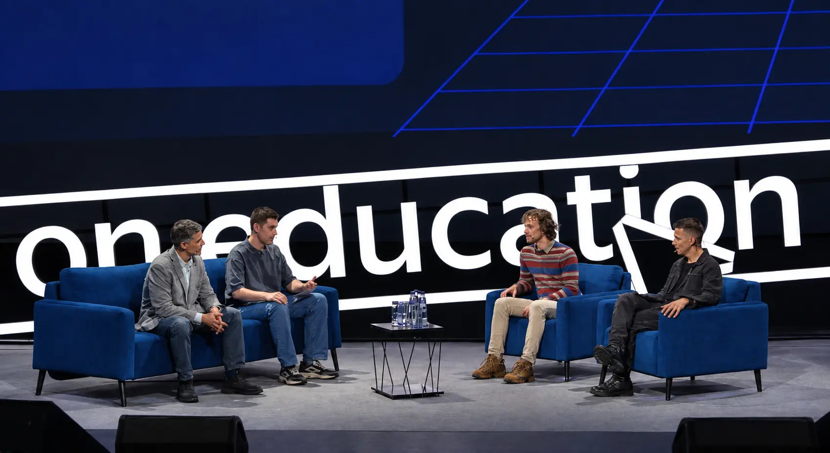 Четыре спикера сидят на синих диванах на сцене конференции oneeducation.