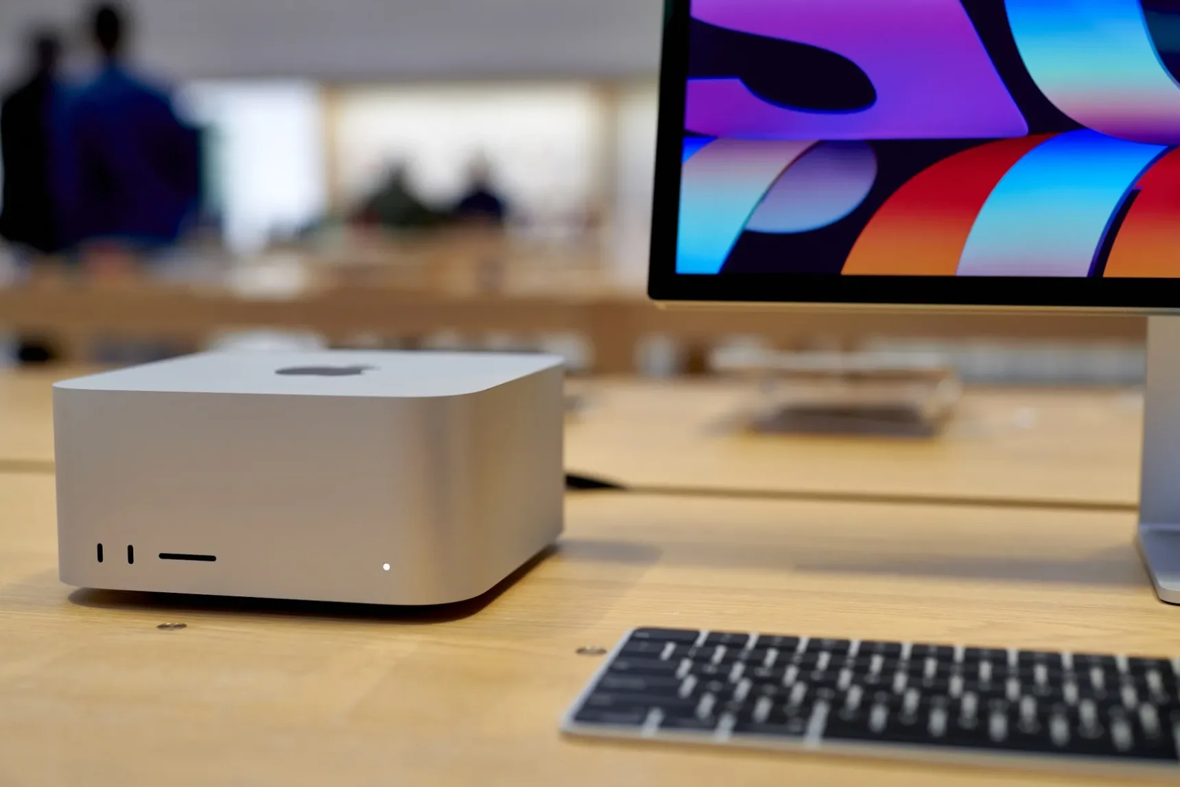 Серебристый Mac Studio, клавиатура и монитор на деревянном столе.