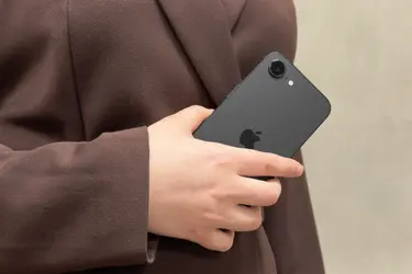 Человек в коричневом пальто держит в руке черный смартфон Apple с логотипом яблока на задней панели.