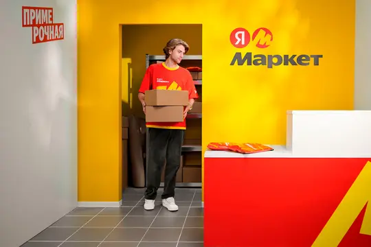 Мужчина в красной футболке Яндекс Маркет держит коробки у желтой стены с логотипом.
