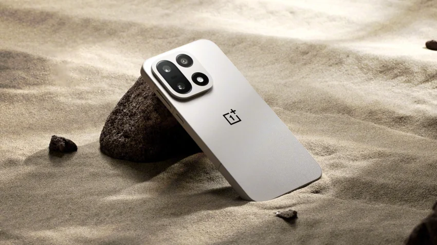 Белый смартфон OnePlus с тройной камерой лежит на песке, опираясь на камень.