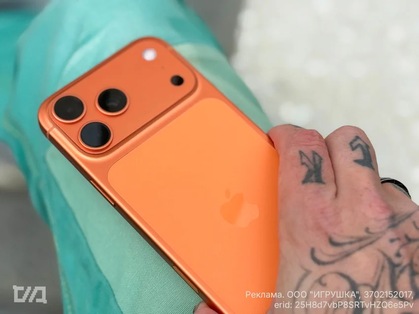 Оранжевый смартфон с логотипом Apple и тройной камерой в руке с татуировками.