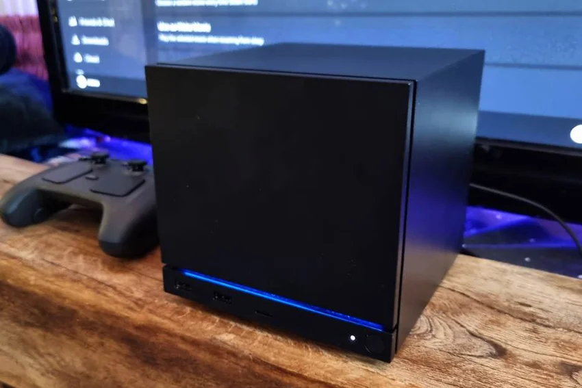 Компактный игровой компьютер Steam Machine рядом с контроллером и монитором с интерфейсом SteamOS.
